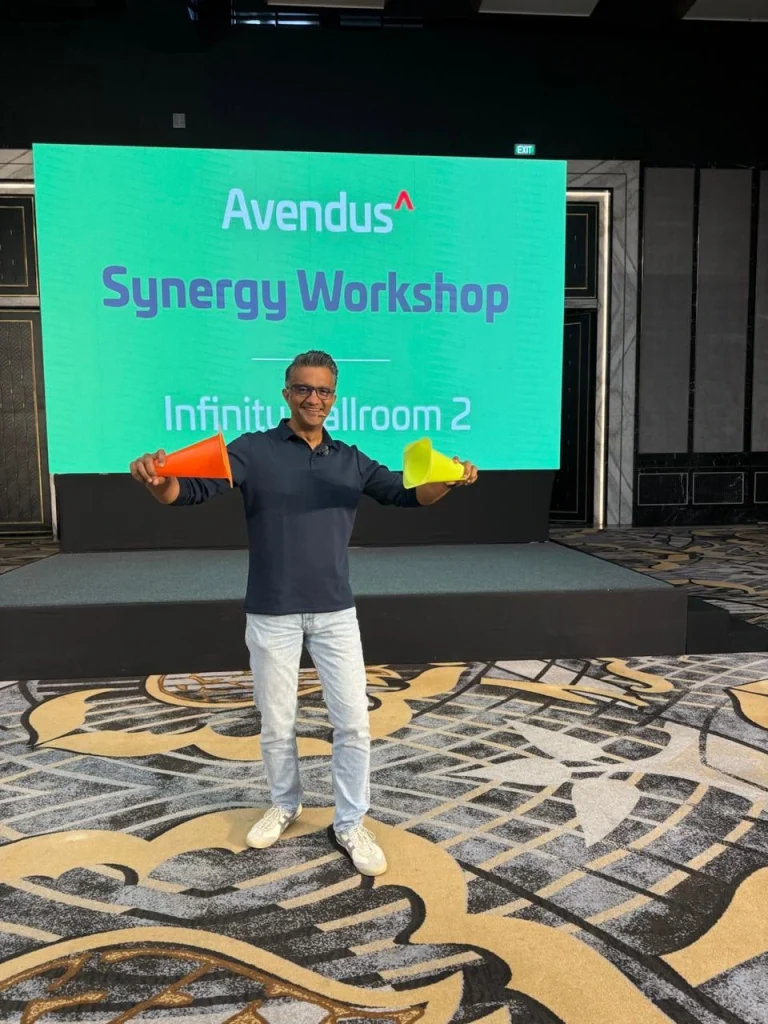 A professional facilitator holding colorful cones on a stage in front of a digital screen titled "Avendus Synergy Workshop" in a ballroom.