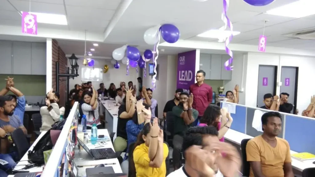 Employees participating in a desk yoga session at a decorated office with purple and white balloons for International Yoga Day.