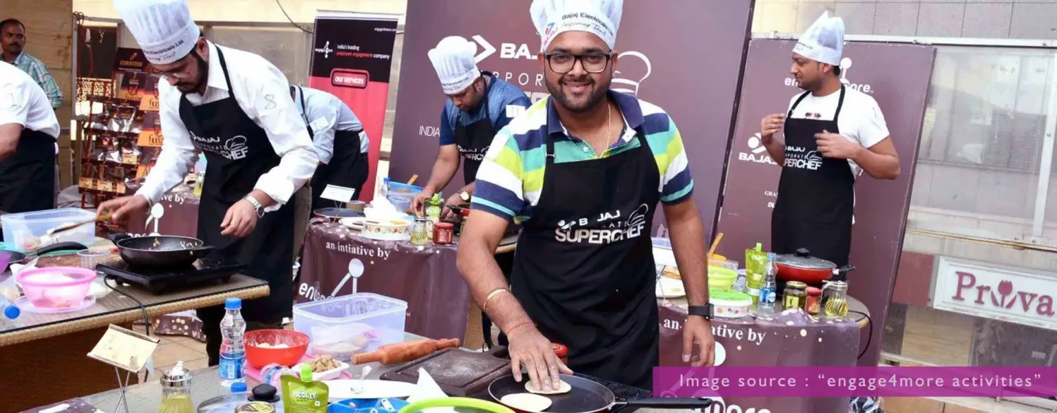 A group of corporate employees wearing chef hats and aprons, actively cooking at individual stations during a "Bajaj Corporate SuperChef" competition outdoors.
