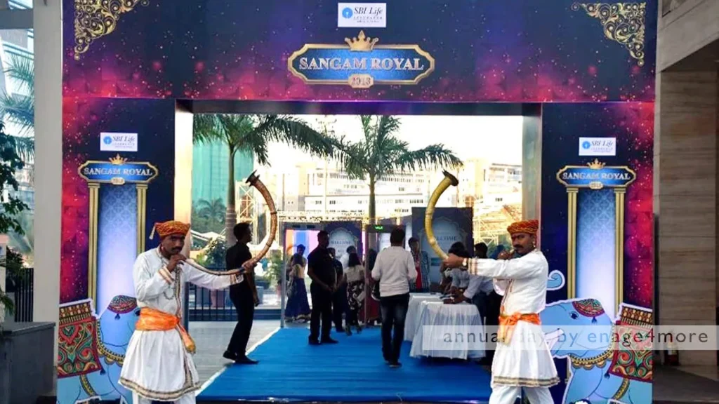 Two men in traditional white and orange attire blowing long brass trumpets at the "Sangam Royal" event entrance gate.