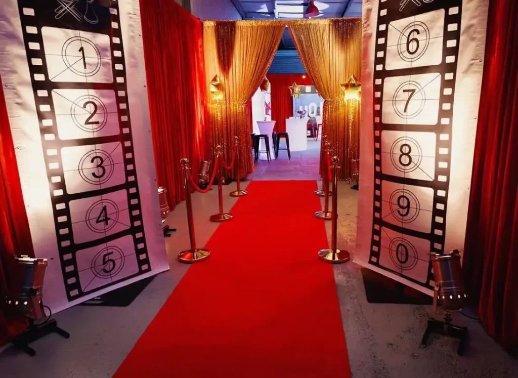 A grand Hollywood-themed entrance featuring a red carpet, gold stanchions, gold sequin curtains, and large filmstrip banners numbered 1 to 0.