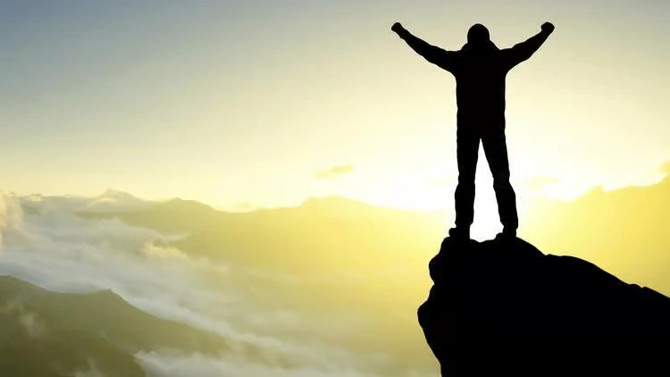 Silhouette of a mountaineer standing with arms raised in triumph at the peak of a mountain representing corporate leadership success