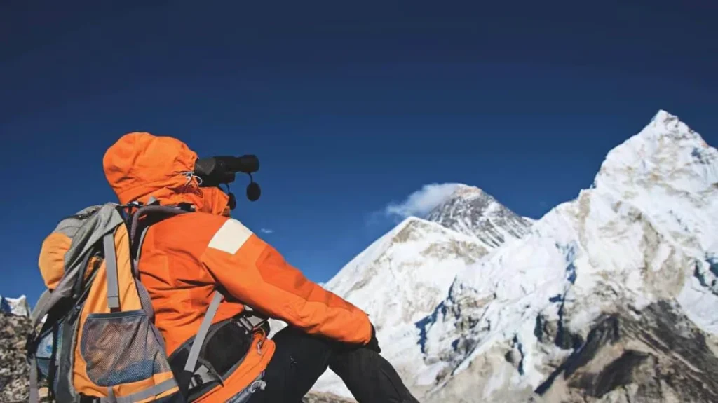 A mountaineer in orange gear looking through binoculars at a distant, snow-capped peak, representing the importance of detailed assessment and preparation in business