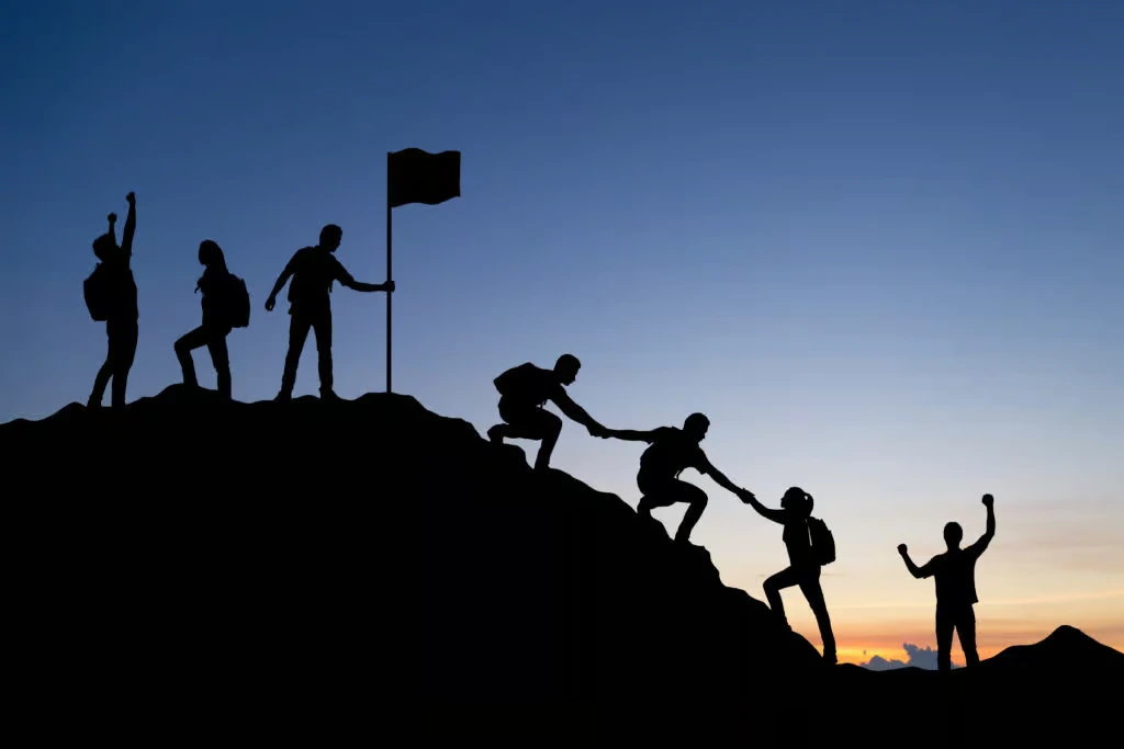 Silhouette of a corporate team helping each other climb a mountain to reach a flag at the summit, symbolizing team synergy and business success