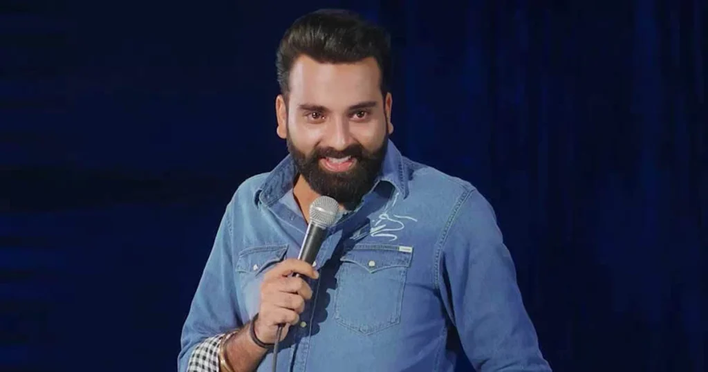 Anubhav Singh Bassi performing on stage, holding a microphone, wearing a denim shirt, with a expressive smile against a dark blue background