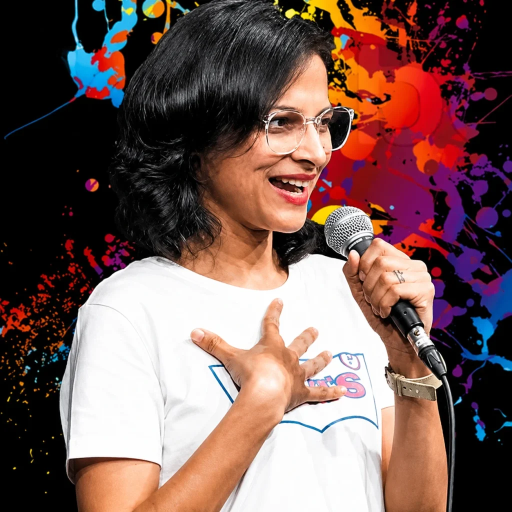 Comedian Rupali Tyagi performing on stage in a white t-shirt, smiling with her hand on her chest, against a vibrant, multi-colored paint splatter background
