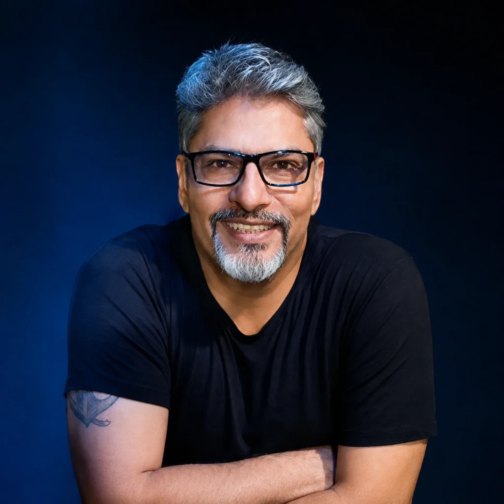 Comedian Manish Tyagi with a friendly smile, wearing glasses and a black t-shirt against a dark blue background