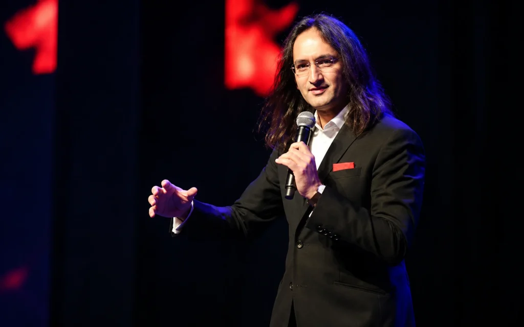 Comedian Papa CJ performing on stage in a suit, holding a microphone and gesturing to the audience