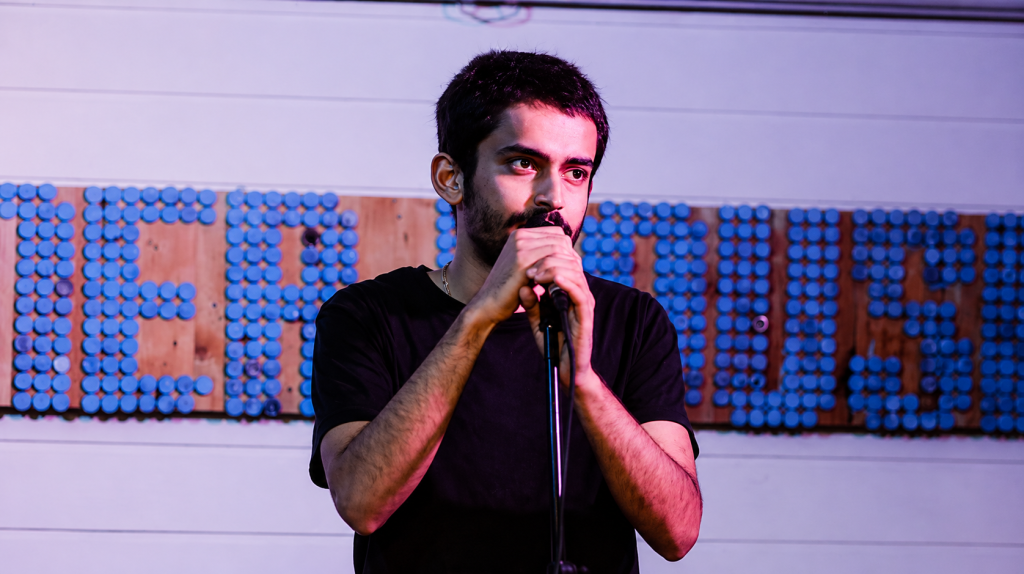 Comedian Shashwat Maheshwari performing stand-up on stage with a microphone in front of a textured backdrop