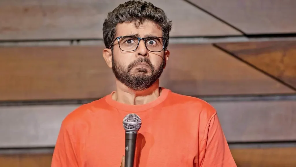 Comedian Aakash Mehta with glasses and a beard, wearing an orange t-shirt, holding a microphone while performing on stage