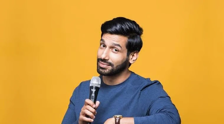 Comedian Kanan Gill smiling while holding a microphone against a plain yellow background