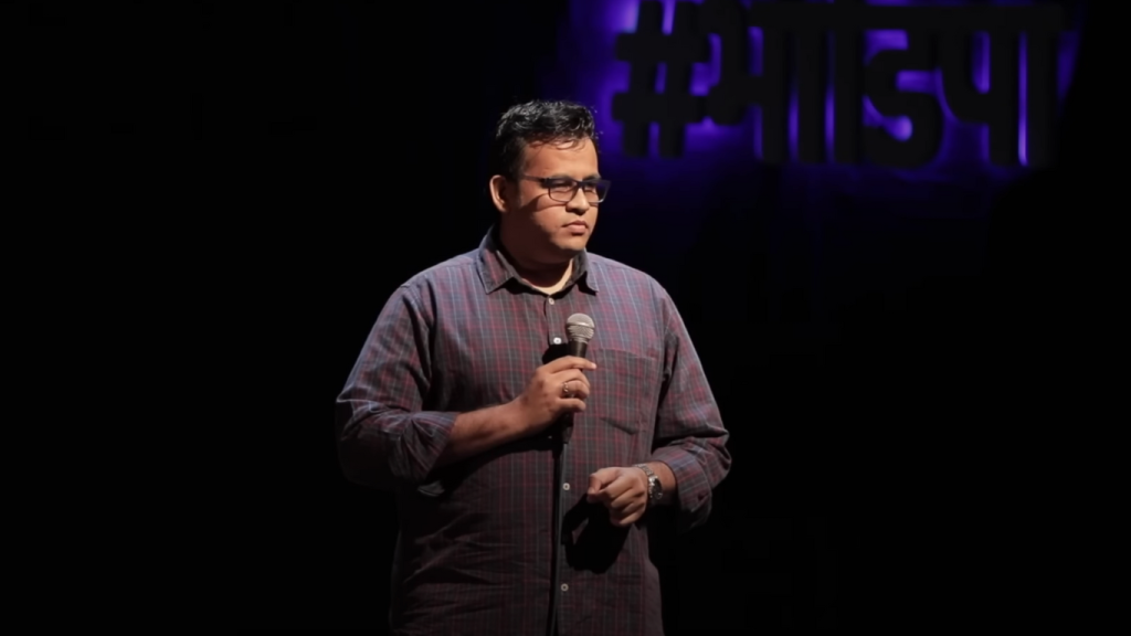 Comedian Mandar Bhide standing on a dark stage holding a microphone, wearing a plaid shirt and glasses