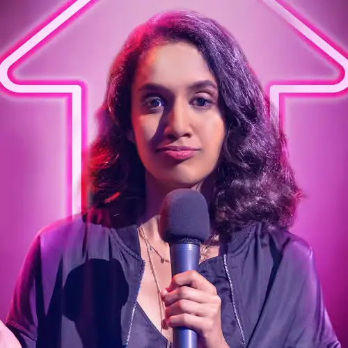 Comedian Niveditha Prakasam performing stand-up comedy on stage, holding a microphone in front of a glowing pink arrow backdrop