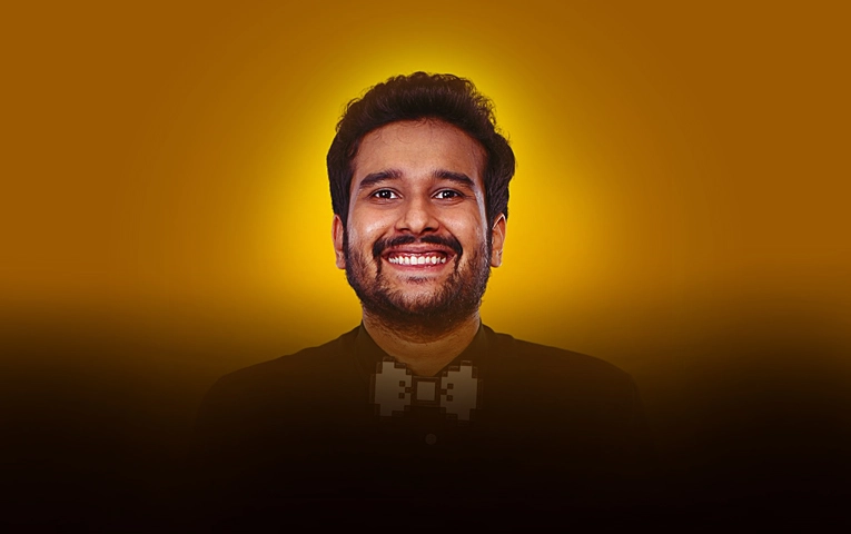 A portrait of Indian comedian Sahil Shah smiling, wearing a dark shirt and a unique pixelated-style bow tie against a warm yellow gradient background