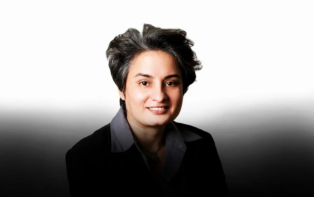 A professional headshot of comedian and activist Vasu Primlani smiling, wearing a dark blazer and collared shirt against a white-to-gray gradient background