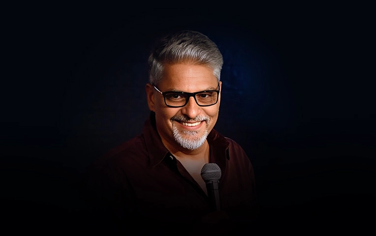 Comedian Commander Manish Tyagi smiling, wearing glasses and a dark shirt, holding a microphone against a dark background