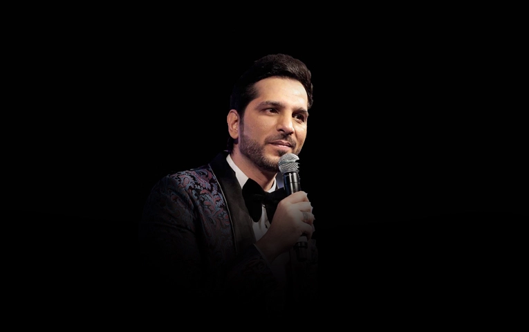 Nitinn R Miranni, also known as Mr. Komic Sutra, performing on stage with a microphone, wearing a patterned tuxedo jacket and bow tie against a black background