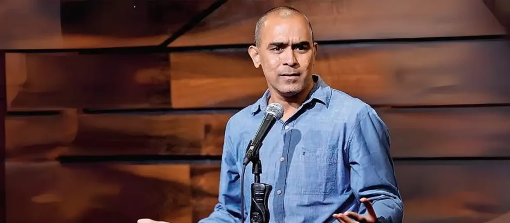 Comedian RajaSekhar Mamidanna performing on stage with a microphone, wearing a blue button-down shirt against a wood-paneled background