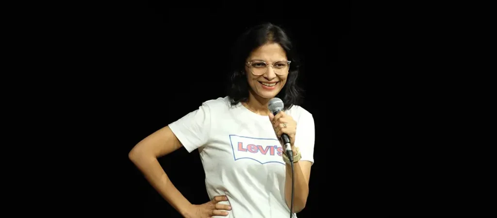 Comedian Rupali Tyagi performing on stage, smiling and holding a microphone, wearing a white t-shirt and glasses