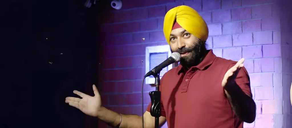 Comedian Vikramjit Singh wearing a yellow turban and a maroon polo shirt, performing on stage with a microphone