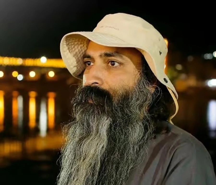 A close-up portrait of Sri Anish wearing a bucket hat, looking off into the distance with a thoughtful expression against a blurred, warm-lit background