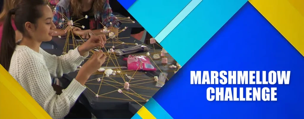 A young woman in a white sweater participates in the Marshmallow Challenge, carefully building a structure using spaghetti sticks and marshmallows on a table.