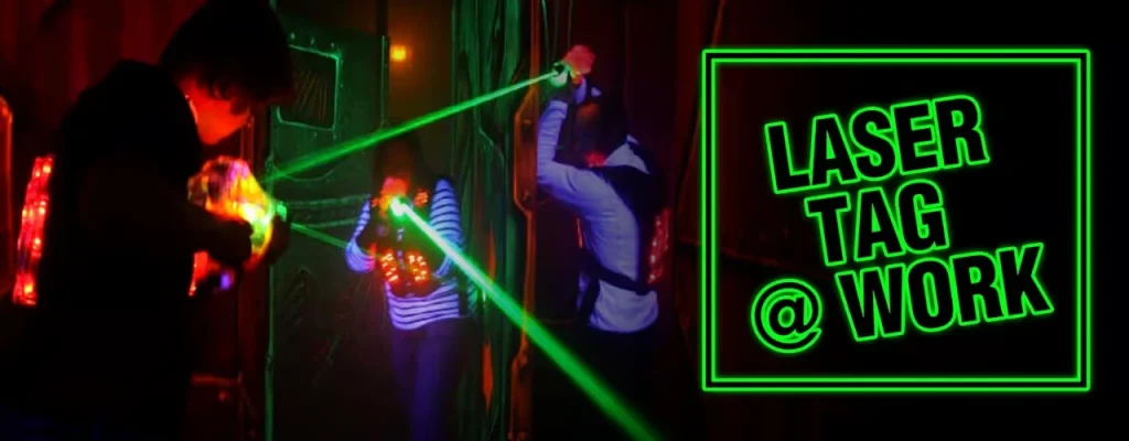 A group of corporate employees wearing glowing vests and using laser guns to play laser tag in a dark, neon-lit indoor arena with a "Laser Tag @ Work" text overlay.
