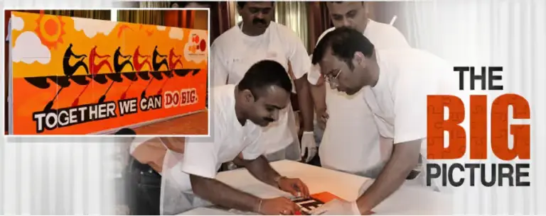 The-Big-Picture-Collaborative-Corporate-Art-Workshop Corporate professionals in white t-shirts working together to assemble a large, colorful mural titled "Together We Can Do Big," featuring a rowing team silhouette.