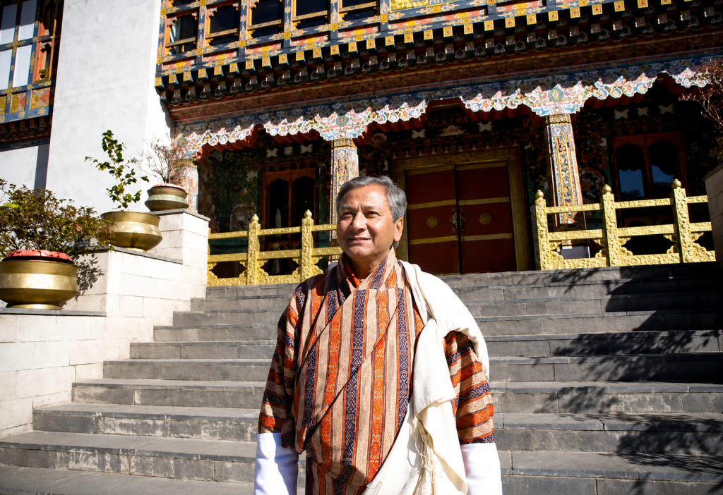 Dr. Saamdu Chetri standing on the steps of a traditional Bhutanese building, wearing a Gho