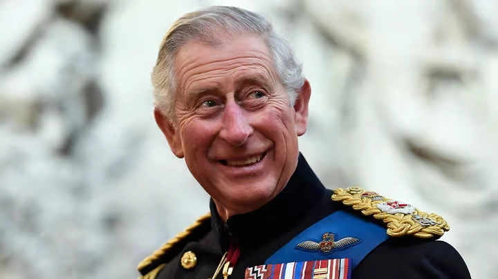 A close-up portrait of King Charles III smiling warmly, dressed in a traditional dark military uniform adorned with gold epaulettes and various service medals.