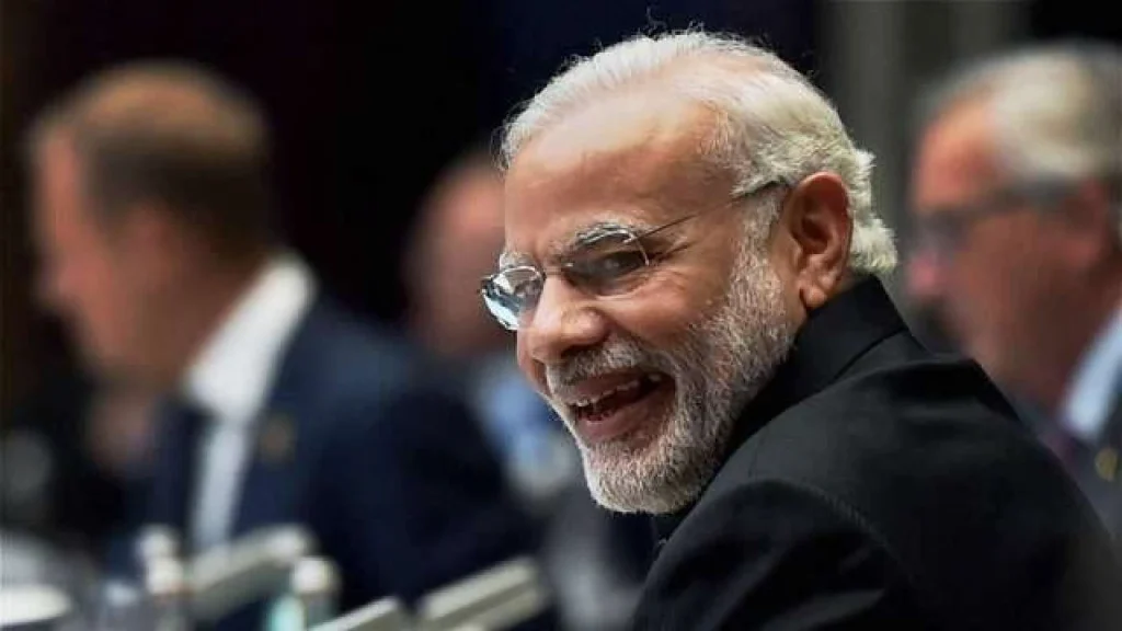 A close-up profile of Prime Minister Narendra Modi smiling and wearing spectacles, set against a blurred background of a formal international summit or meeting.