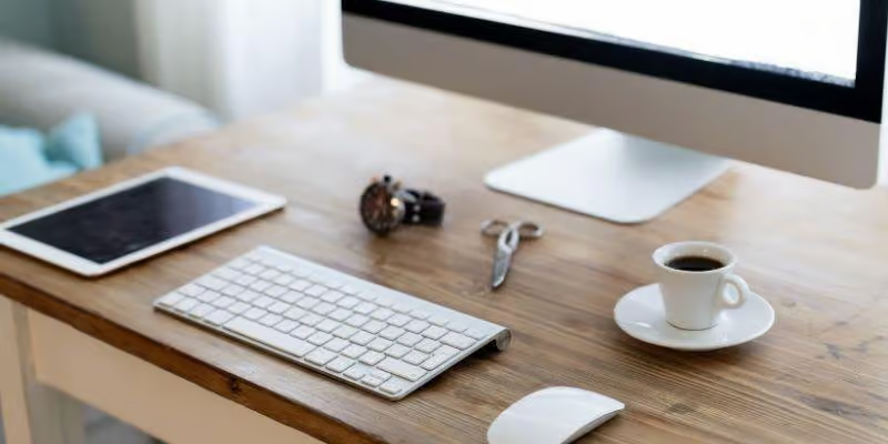 Office-Branding-Elements-Desktop-Setup A minimalist, high-angle view of a clean wooden office desk featuring a white keyboard, a mouse, a digital tablet, a pair of scissors, a watch, and a white cup of coffee on a saucer.