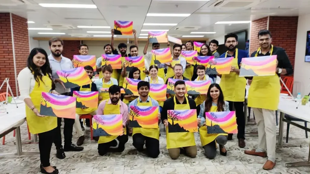 A large group of corporate employees wearing yellow aprons in a modern office, each holding up a colorful sunset painting they created during a team-building workshop.