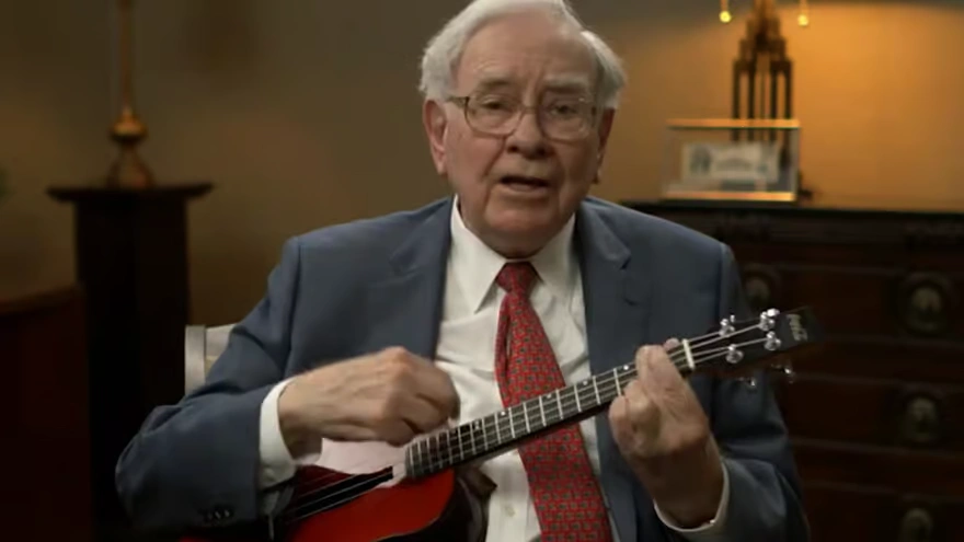 A candid shot of Warren Buffett in a blue suit and red tie, smiling while playing a small red ukulele in a cozy, indoor setting.