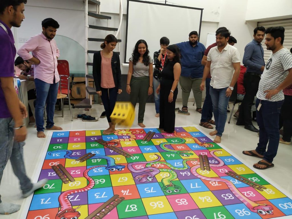 A group of young corporate professionals playing a life-sized version of Snakes and Ladders on an office floor, with a large yellow foam die in mid-air.
