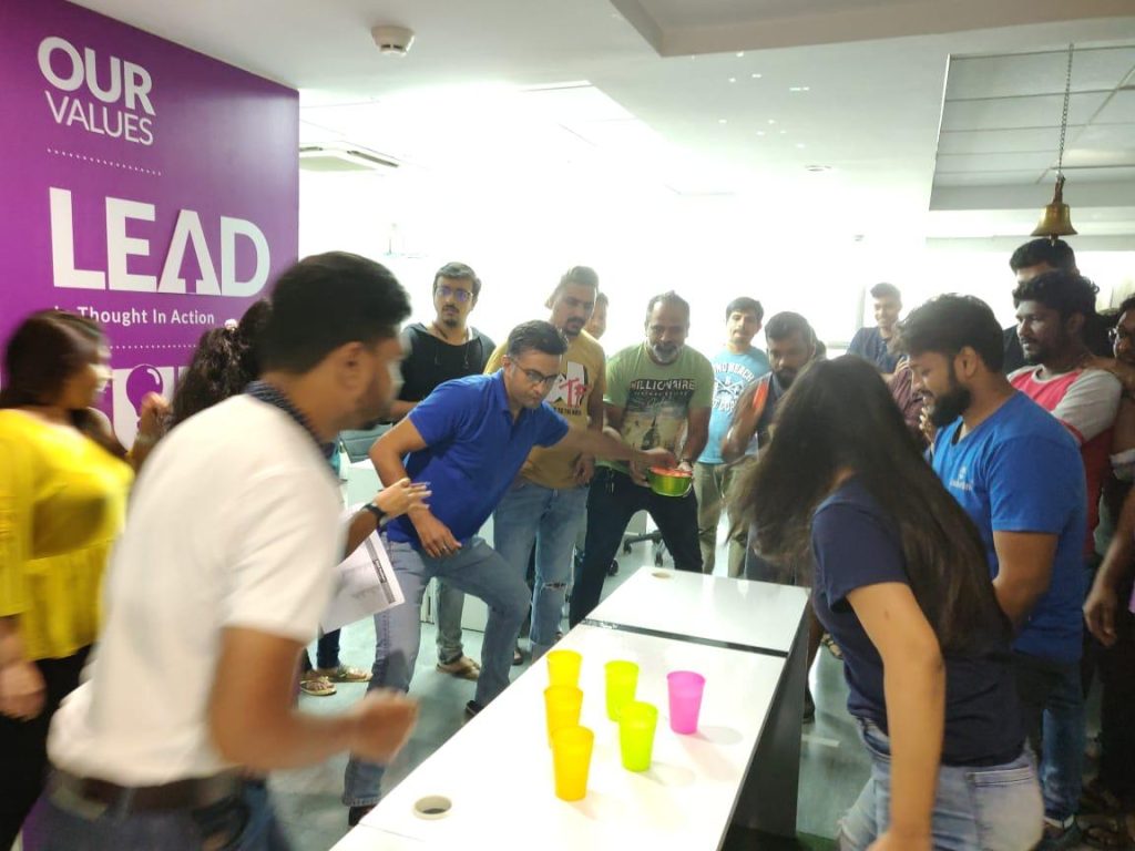 A group of corporate employees in a high-energy office setting participating in a competitive cup-stacking activity under a wall that reads "Our Values: LEAD Thought In Action."
