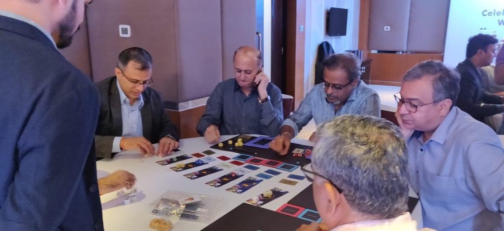 A group of male corporate professionals in formal and semi-formal attire sitting around a table, intensely focused on a strategic board game simulation during a workshop.