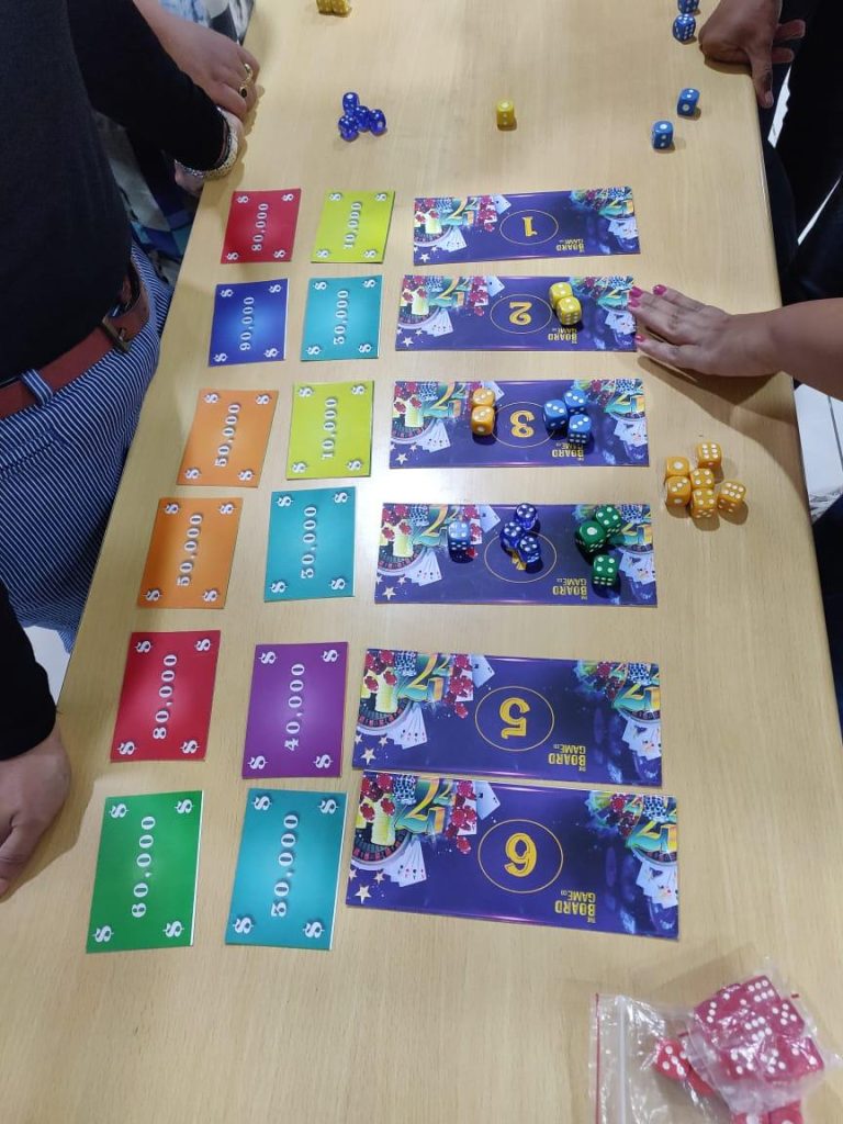 Top-down view of a corporate team-building board game with numbered mats, colored currency cards (10,000 to 80,000), and various dice being used by participants.