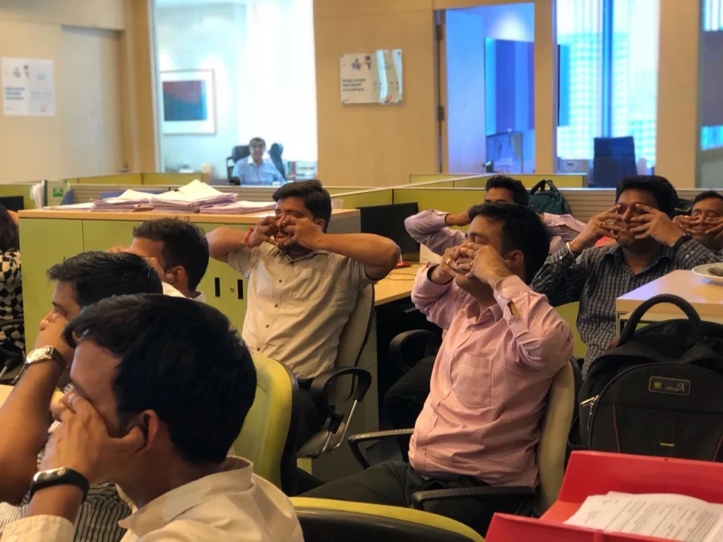A group of Indian male corporate employees in formal shirts sitting in their office cubicles and practicing a "Shanmukhi Mudra" yoga posture by placing their fingers over their eyes and face for a mindfulness session.