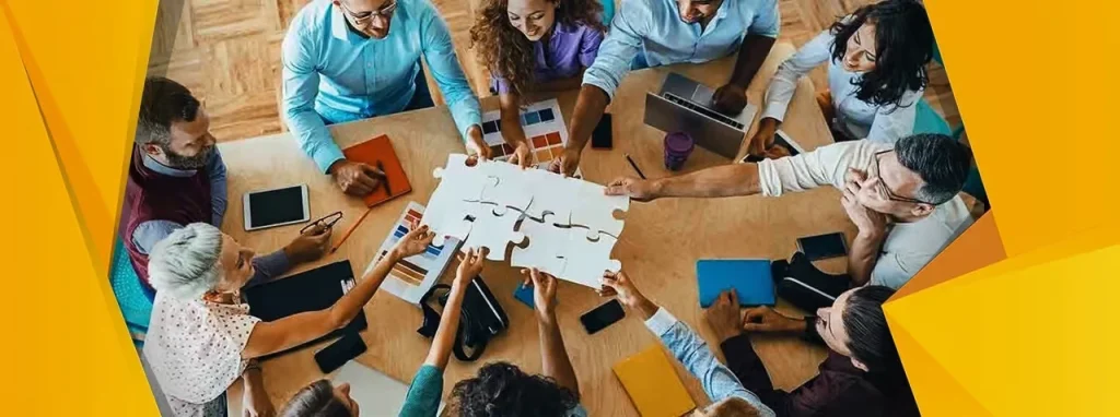 An overhead view of a diverse group of corporate professionals sitting around a wooden table, working together to fit large white jigsaw puzzle pieces together in a modern office environment.