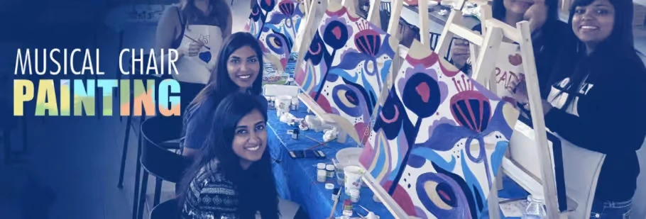 Four female Indian professionals smiling while working on vibrant, abstract floral paintings on easels during a "Musical Chair Painting" session in a modern office.