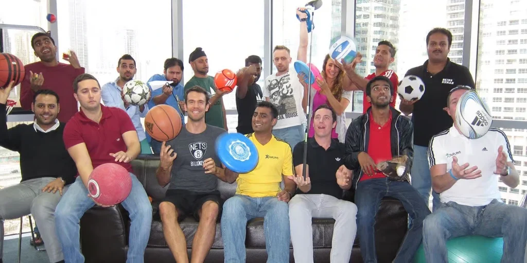 A group of approximately fifteen diverse corporate professionals in an office setting, posing together while playfully holding various sports equipment including basketballs, footballs, frisbees, a golf club, and a tennis racket.