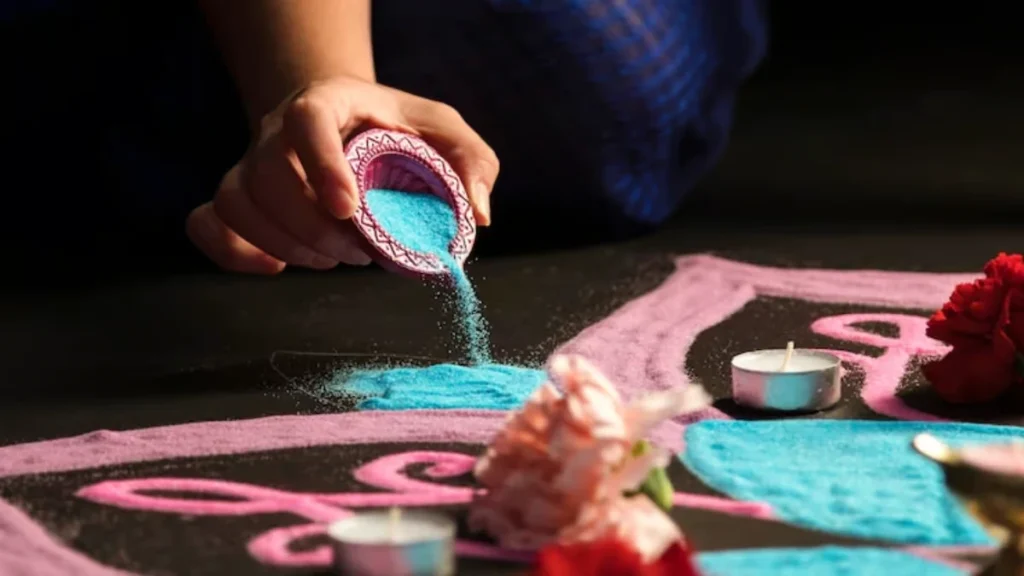 A close-up shot of an employee's hand carefully pouring bright blue decorative powder from a small purple clay diya to create an intricate Rangoli pattern on an office floor, surrounded by pink powder borders, lit candles, and pink flowers.