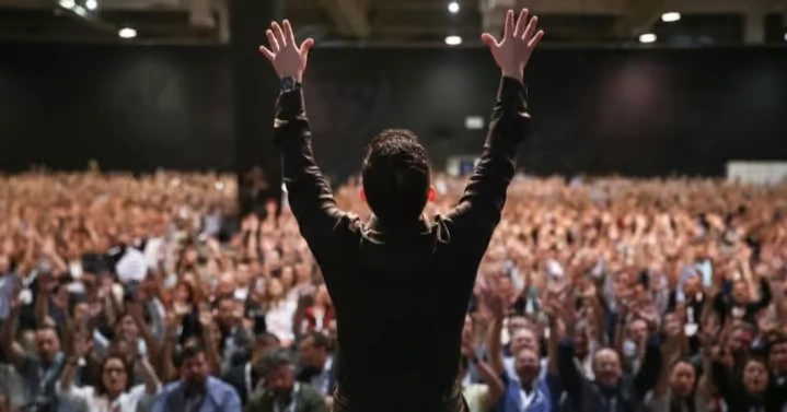 A back view of a leader standing on stage with arms raised toward a massive, cheering crowd of corporate professionals in a large convention hall, symbolizing unified organizational purpose.