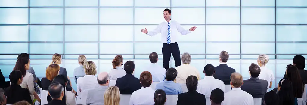 A professional male motivational speaker in a white shirt and blue tie standing on a stage, gesturing with open arms while addressing an attentive corporate audience in a modern, brightly lit hall with a grid-patterned glass wall.