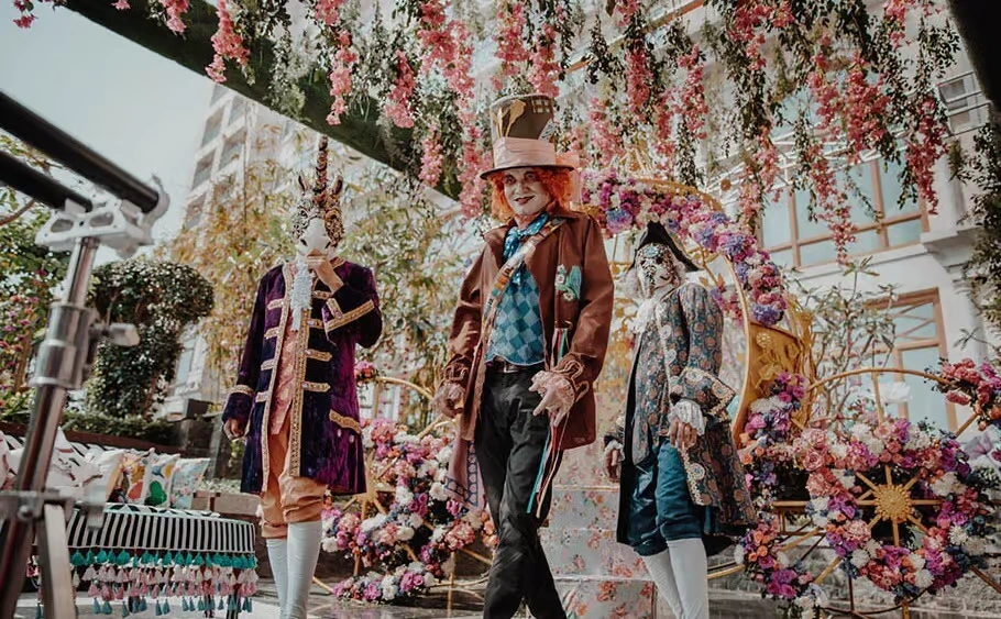 Three performers in elaborate Alice in Wonderland costumes, including the Mad Hatter and a figure in a unicorn mask, posing in front of a floral-covered carriage in an outdoor setting.