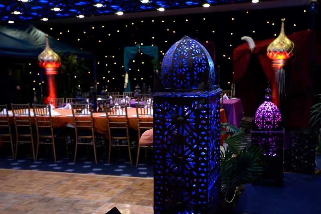 A dimly lit corporate event space decorated in an Arabian Nights theme, featuring ornate blue and purple lanterns, gold-domed structures, and long banquet tables with orange linens under a starlit ceiling.