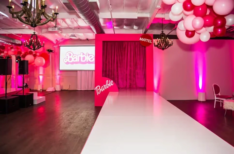 A high-end corporate event space decorated in a Barbie theme, featuring a long white runway, a large pink "Barbie" branded box entrance, pink lighting, and pink and white balloon installations.
