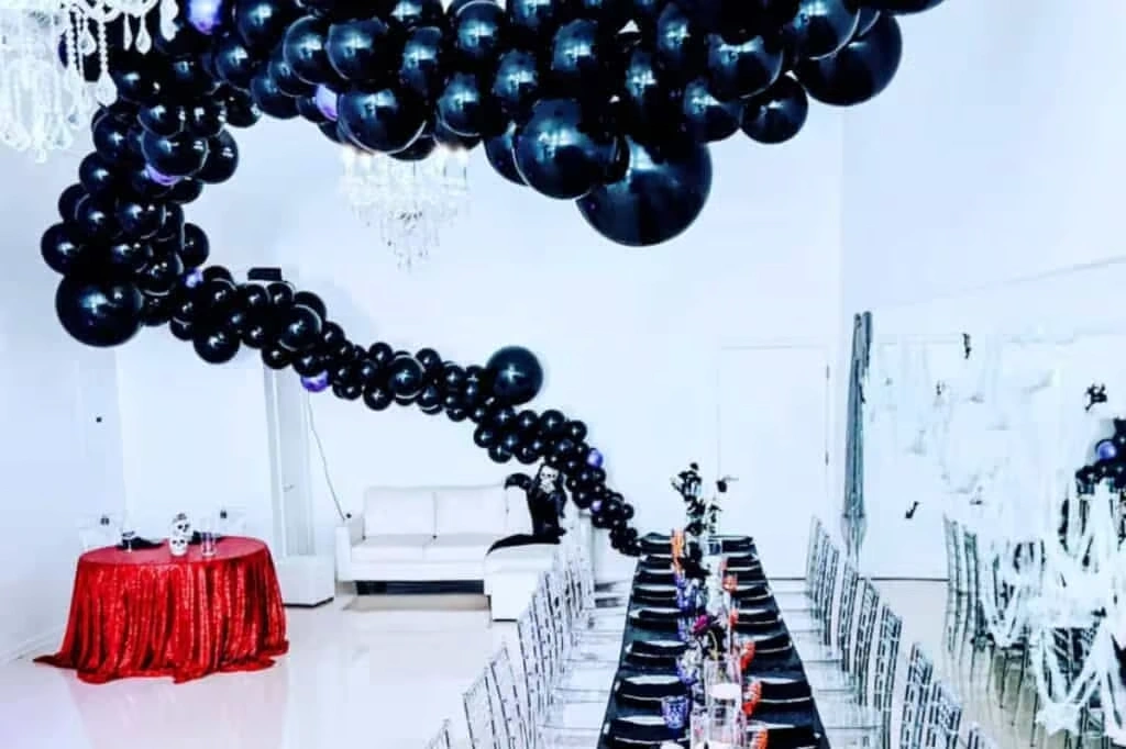 A bright corporate event room with white walls, featuring a massive, winding black balloon installation stretching across the ceiling, long black dining tables with silver chairs, and a single round table with a shimmering red tablecloth.