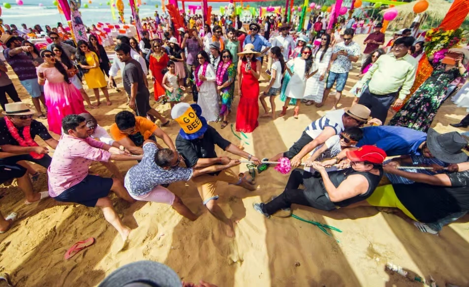 A vibrant, sun-drenched beach carnival scene where two teams are engaged in a competitive tug-of-war on the sand, surrounded by colorful decor, tents, and cheering colleagues.